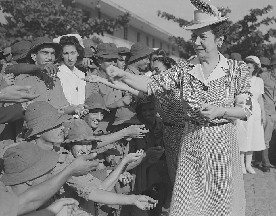 Voluntária da Cruz Vermelha Brasileira, de uniforme, chapéu e braçadeira da cruz vermelha no braço esquerdo, entrega itens a soldados da FEB, com enfermeiras de branco ao fundo
