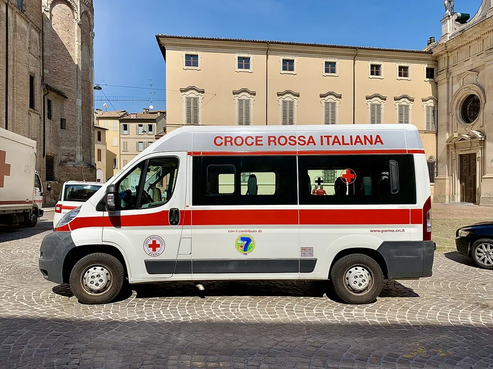 Ambulância branca e vermelha da Cruz Vermelha Italiana numa praça de Parma, com o nome em letras grandes e o emblema pequeno