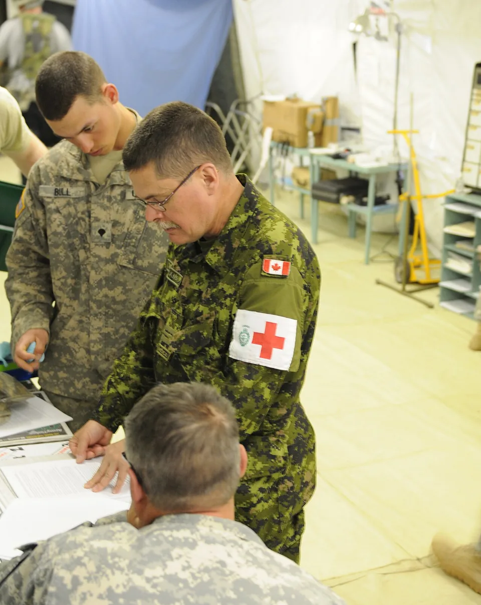 Oficial enfermeiro militar canadense, de uniforme camuflado e braçadeira branca com a cruz vermelha, orientando dois soldados numa tenda de hospital de campanha