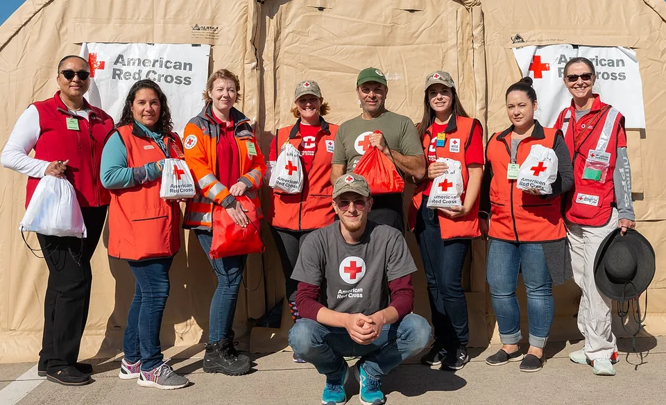 Voluntários da Cruz Vermelha Americana de coletes vermelhos com o emblema, posando sorrindo diante de uma tenda com cartazes da organização
