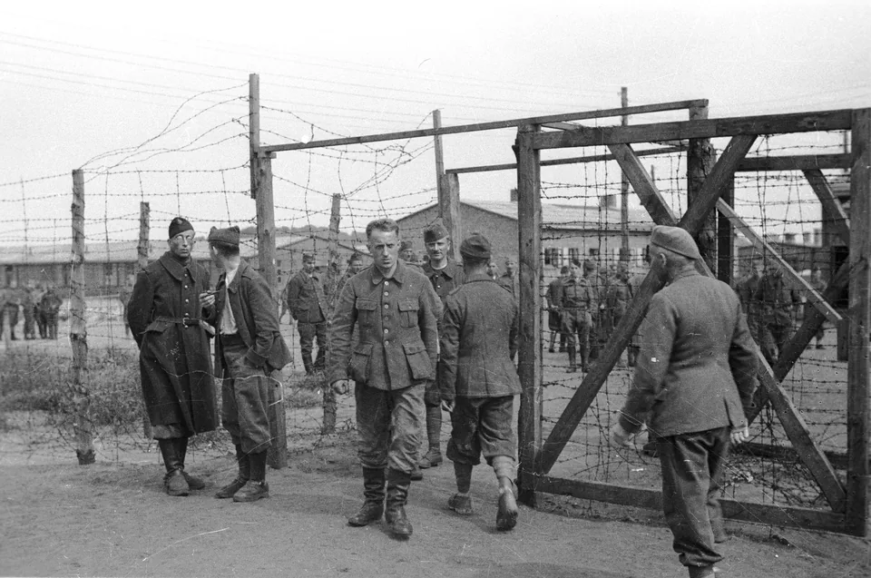 Portão de madeira e arame farpado do campo de prisioneiros Stalag X B, em Sandbostel, com prisioneiros de uniforme conversando junto à cerca e barracões ao fundo