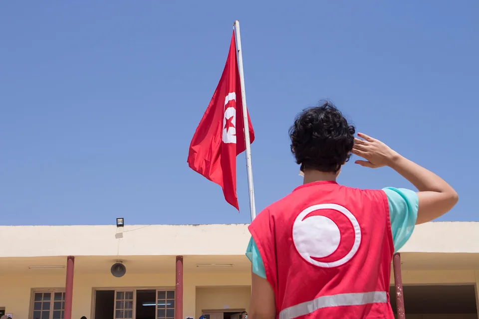 Pessoa voluntária do Crescente Vermelho Tunisiano, de costas, com colete vermelho com o crescente branco, faz continência à bandeira da Tunísia diante de um prédio baixo