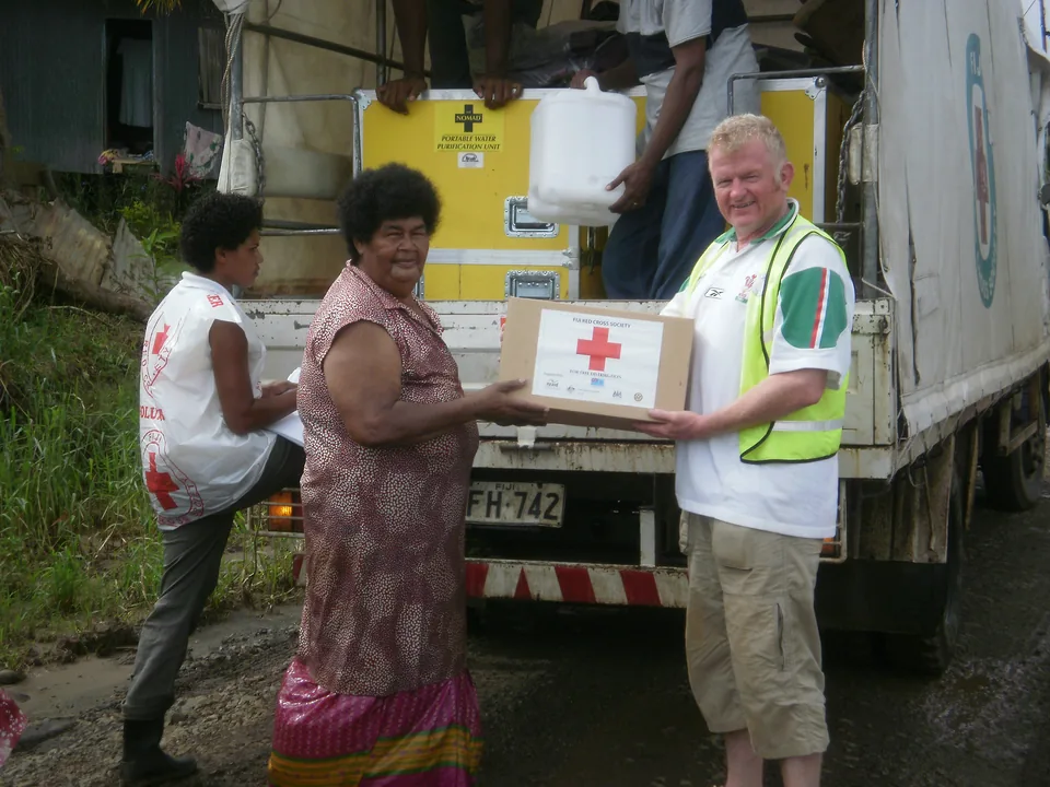 Homem de colete refletivo entrega a uma moradora uma caixa com a cruz vermelha e o nome Fiji Red Cross Society, diante de um caminhão; ao lado, uma voluntária de colete branco com o emblema faz anotações