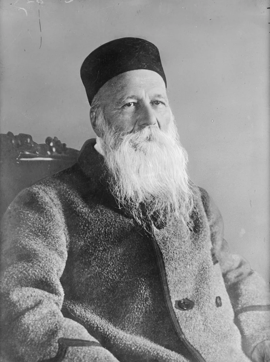 Henri Dunant idoso, sentado numa cadeira de madeira, com longa barba branca, gorro escuro e casaco grosso de lã, olhando para a câmera