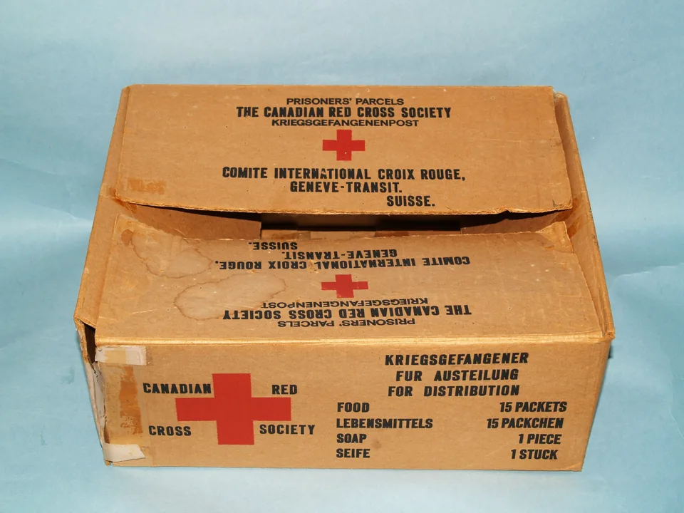 Caixa de papelão da Cruz Vermelha Canadense para prisioneiros de guerra, com a cruz vermelha, a inscrição Prisoners' Parcels e a lista do conteúdo em inglês e alemão