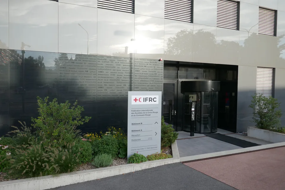 Entrada da sede da Federação Internacional em Genebra: fachada de vidro escuro com os Princípios Fundamentais gravados, porta giratória e totem com o logotipo IFRC