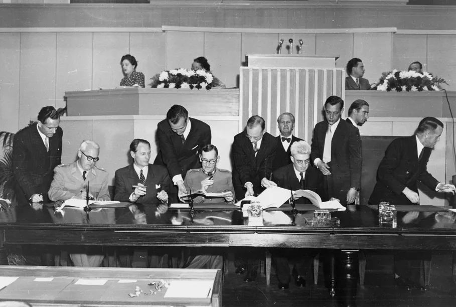Delegados sentados lado a lado a uma longa mesa, assinando e folheando documentos, com assessores em pé atrás, na assinatura das Convenções de Genebra de 1949