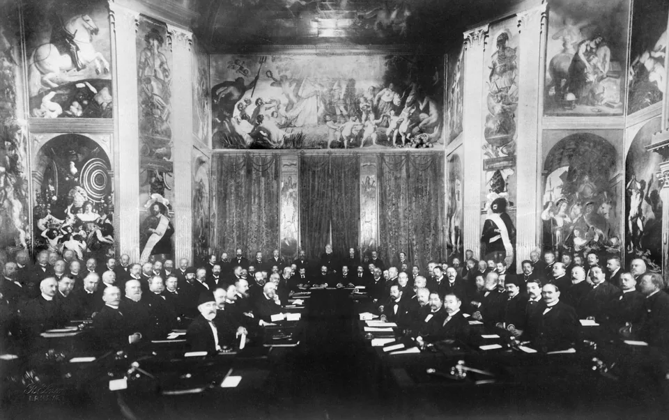 Delegados de terno escuro sentados em mesas dispostas em U, num salão com grandes pinturas murais, na Primeira Conferência Internacional da Paz, em Haia, 1899