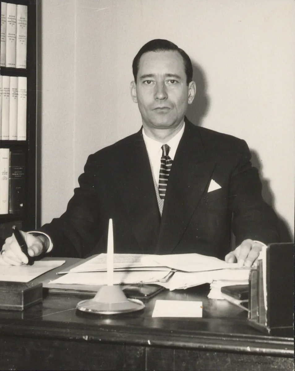 Jean Pictet, de terno escuro e gravata listrada, sentado à mesa de trabalho com uma caneta na mão e estantes de pastas ao fundo, na sede do CICV em Genebra, em 1937
