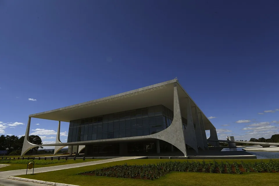 Palácio do Planalto, em Brasília, visto em diagonal: colunas curvas brancas, fachada de vidro, gramado e espelho d'água sob céu azul-escuro