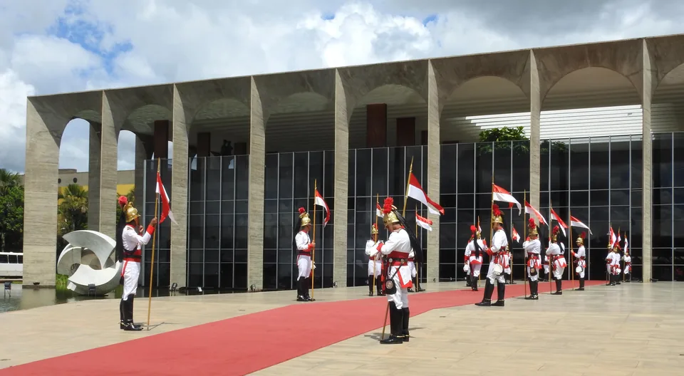 Fachada do Palácio do Itamaraty, em Brasília, com arcos de concreto sobre vidro, espelho d'água com escultura branca e a guarda de honra em uniforme de gala sobre um tapete vermelho
