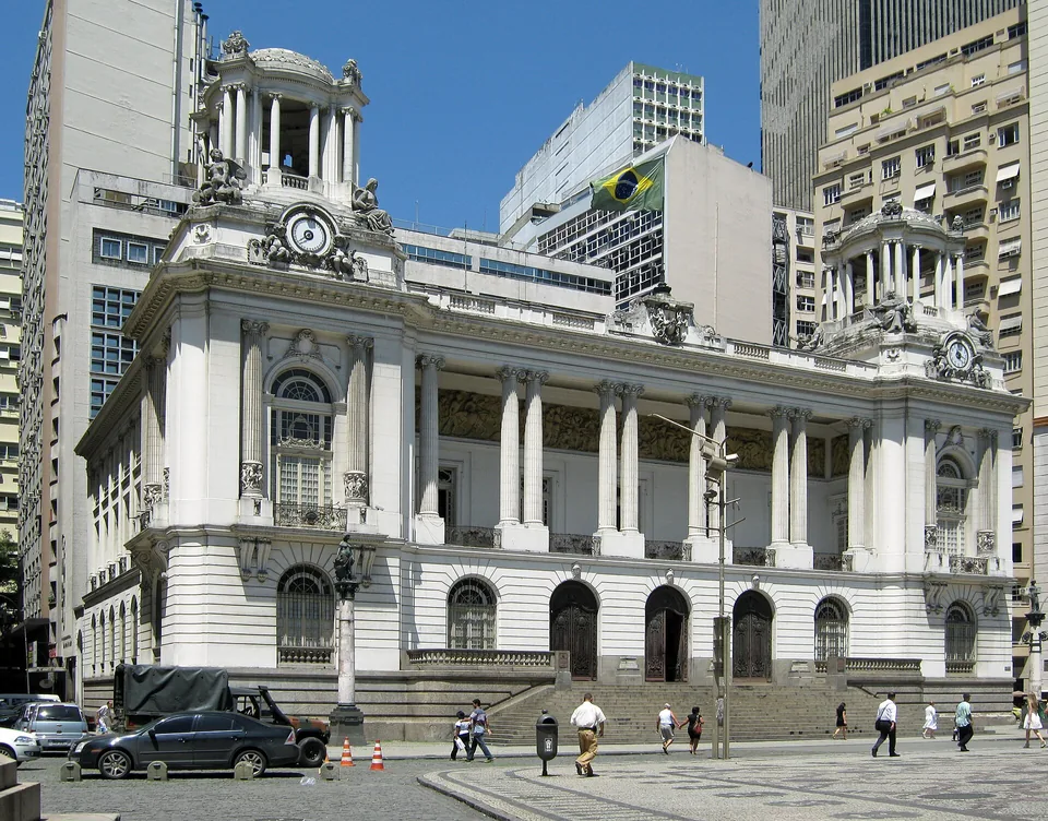Palácio Pedro Ernesto, na Cinelândia: fachada branca eclética com duas torres de relógio e a bandeira do Brasil no mastro, pedestres no calçadão de pedras portuguesas e prédios altos ao fundo