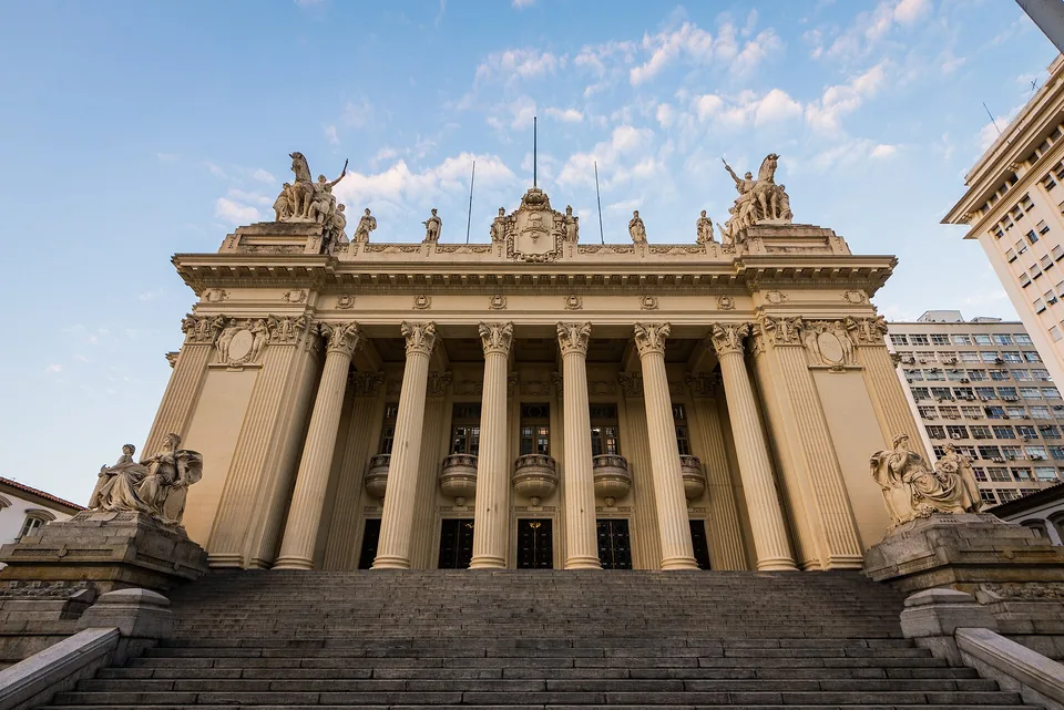 Fachada principal do Palácio Tiradentes, no Centro do Rio: colunata de seis colunas coríntias, frontão com estátuas e duas esculturas sentadas ao lado da escadaria, sob céu azul no fim da tarde
