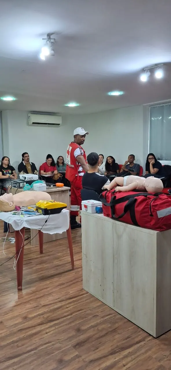 Instrutor da Cruz Vermelha em sala de aula, com manequins e um DEA de treino sobre a mesa