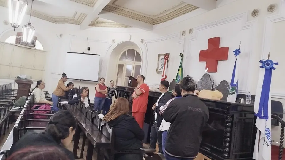 Turma sentada no salão da sede, com a cruz vermelha ao fundo e um manequim sobre a mesa