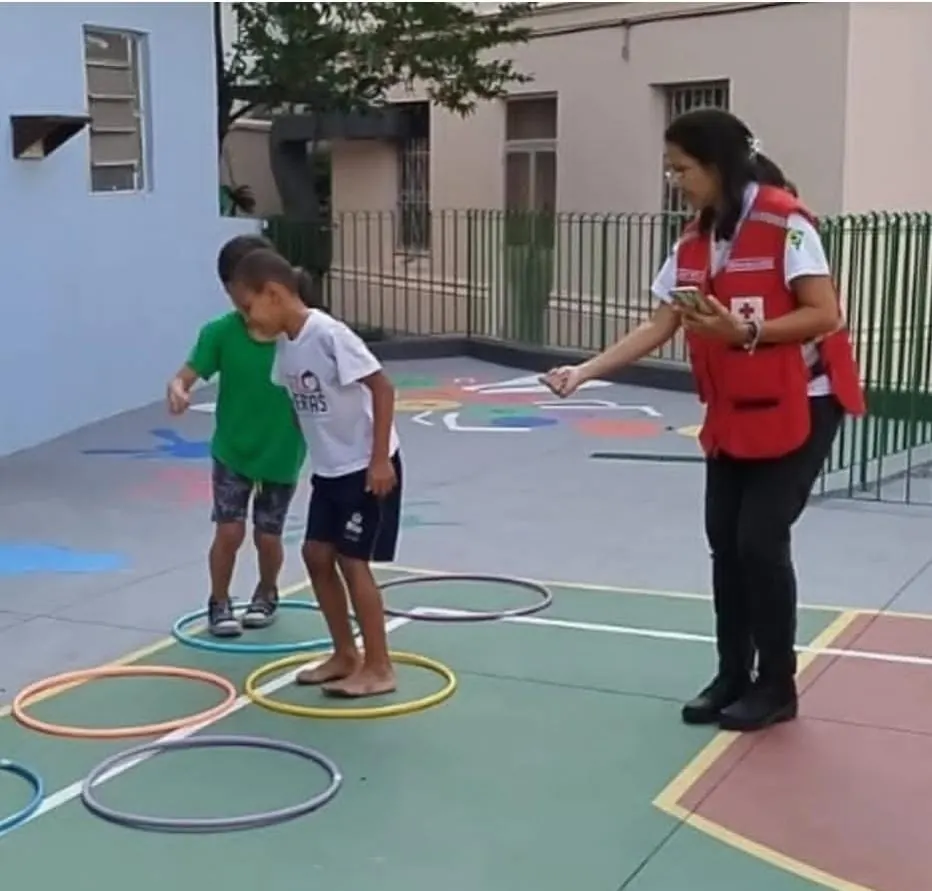 Voluntária da filial, de colete vermelho, conduz dois meninos numa brincadeira de pular dentro de arcos coloridos numa quadra