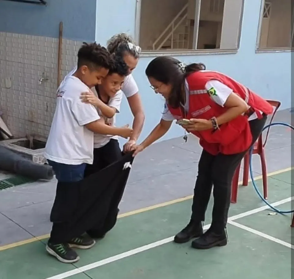 Voluntária da Cruz Vermelha, de colete vermelho, ajuda dois meninos a entrar no saco para a corrida, numa quadra