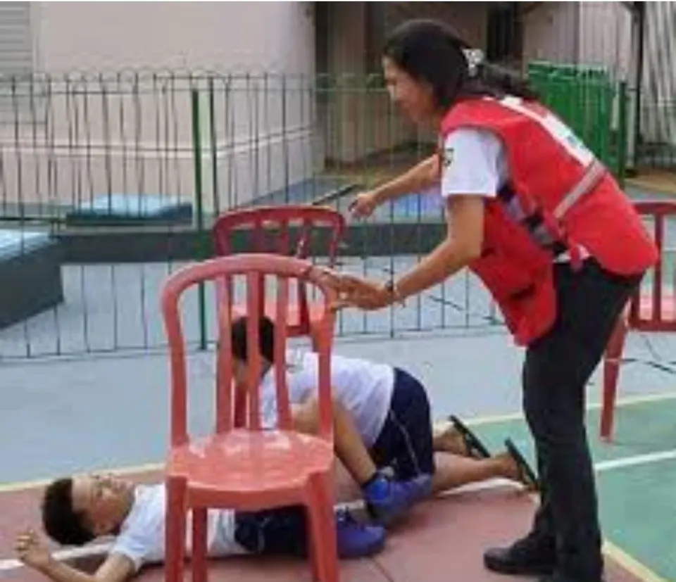 Voluntária da filial, de colete vermelho, orienta jovens numa atividade prática de primeiros socorros, com um deles deitado no chão de uma quadra