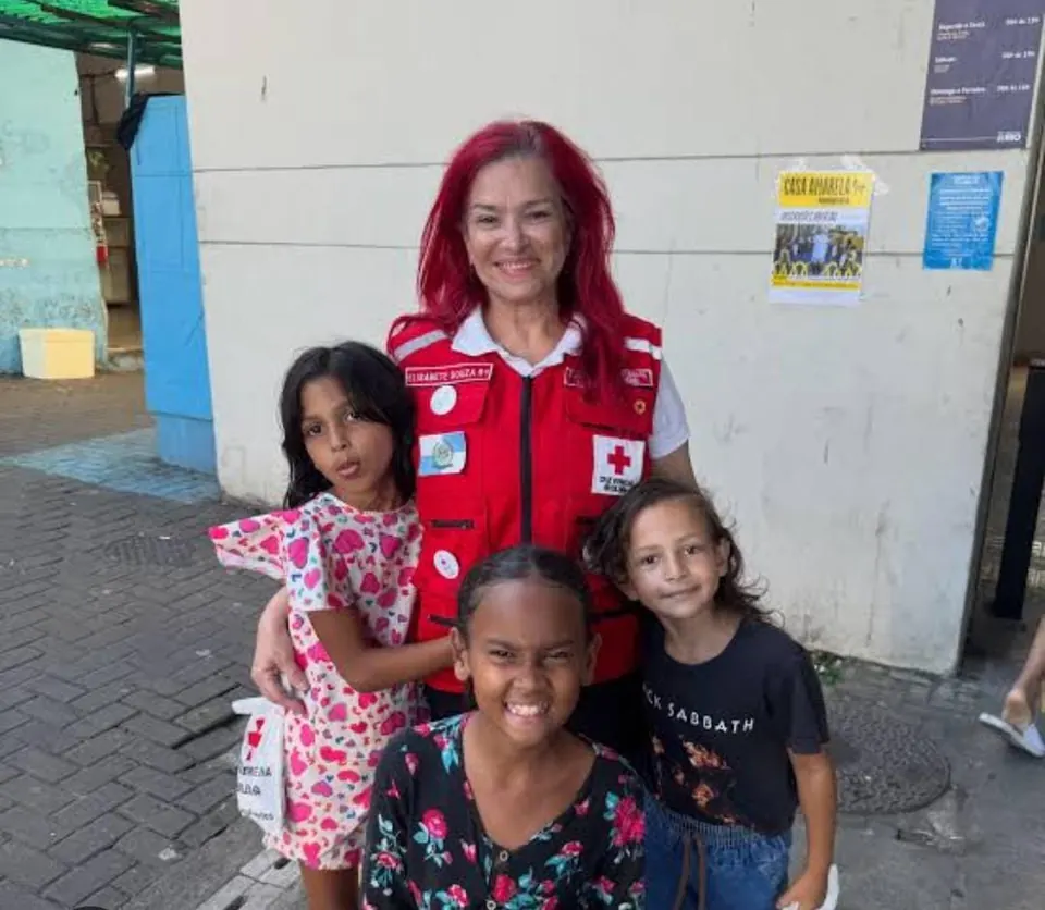 Voluntária da filial, de colete vermelho com o emblema da Cruz Vermelha, sorrindo abraçada a três meninas numa calçada