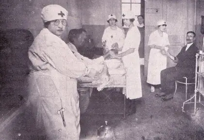 Enfermeiras da Cruz Vermelha Brasileira, de uniforme branco e touca com a cruz, atendem pacientes numa enfermaria; em primeiro plano, uma delas, de óculos, cuida de um paciente deitado numa maca