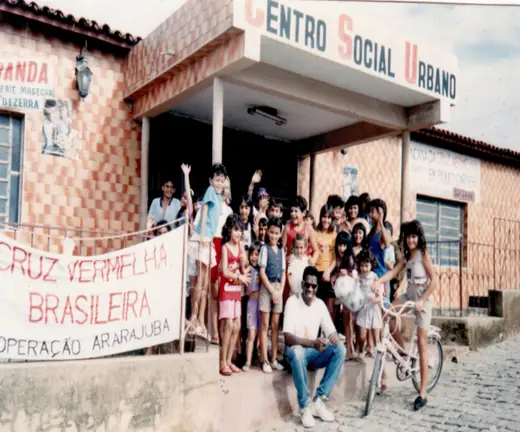 Children and an adult seated at the entrance of an Urban Social Centre, with an Operation Ararajuba banner
