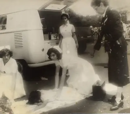Fotografia antiga em preto e branco: duas voluntárias de uniforme branco, ajoelhadas, atendem uma pessoa deitada no chão ao lado de uma Kombi; uma terceira, de touca, acompanha de pé, e uma mulher de roupa escura observa