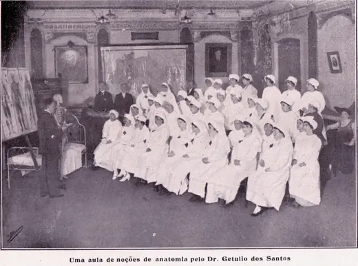 Enfermeiras de uniforme branco e véu, sentadas, assistem a uma aula de anatomia; à esquerda, o professor junto a um esqueleto