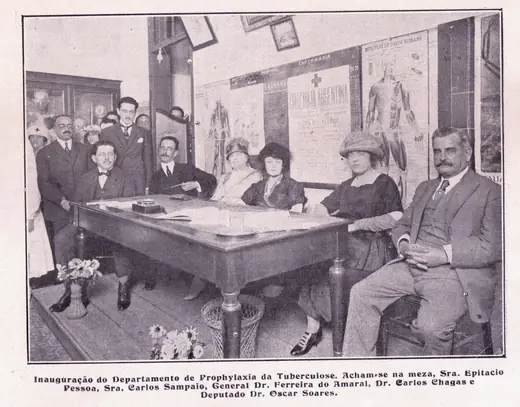 Autoridades e dirigentes sentados em torno de uma mesa, numa sala com cartazes de saúde na parede, na inauguração do Departamento de Profilaxia da Tuberculose