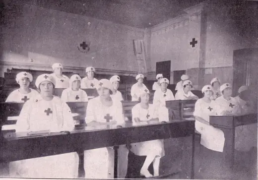 Alunas de uniforme branco com a cruz vermelha, sentadas em carteiras de madeira numa sala de aula