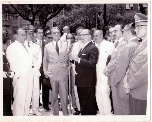 Officials gathered outdoors; a man in a suit speaks in front of a microphone