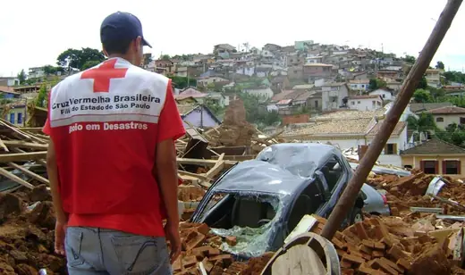 Volunteer seen from behind, in a Brazilian Red Cross vest, looking at destroyed houses and a car on a hillside