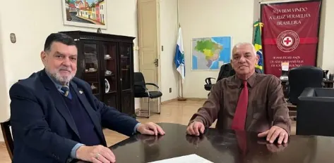 O presidente Luiz Carlos dos Santos e o vice-presidente Antônio Pedregal sentados à mesa do gabinete da presidência, com as bandeiras do Brasil e do estado e a faixa da Cruz Vermelha Brasileira ao fundo