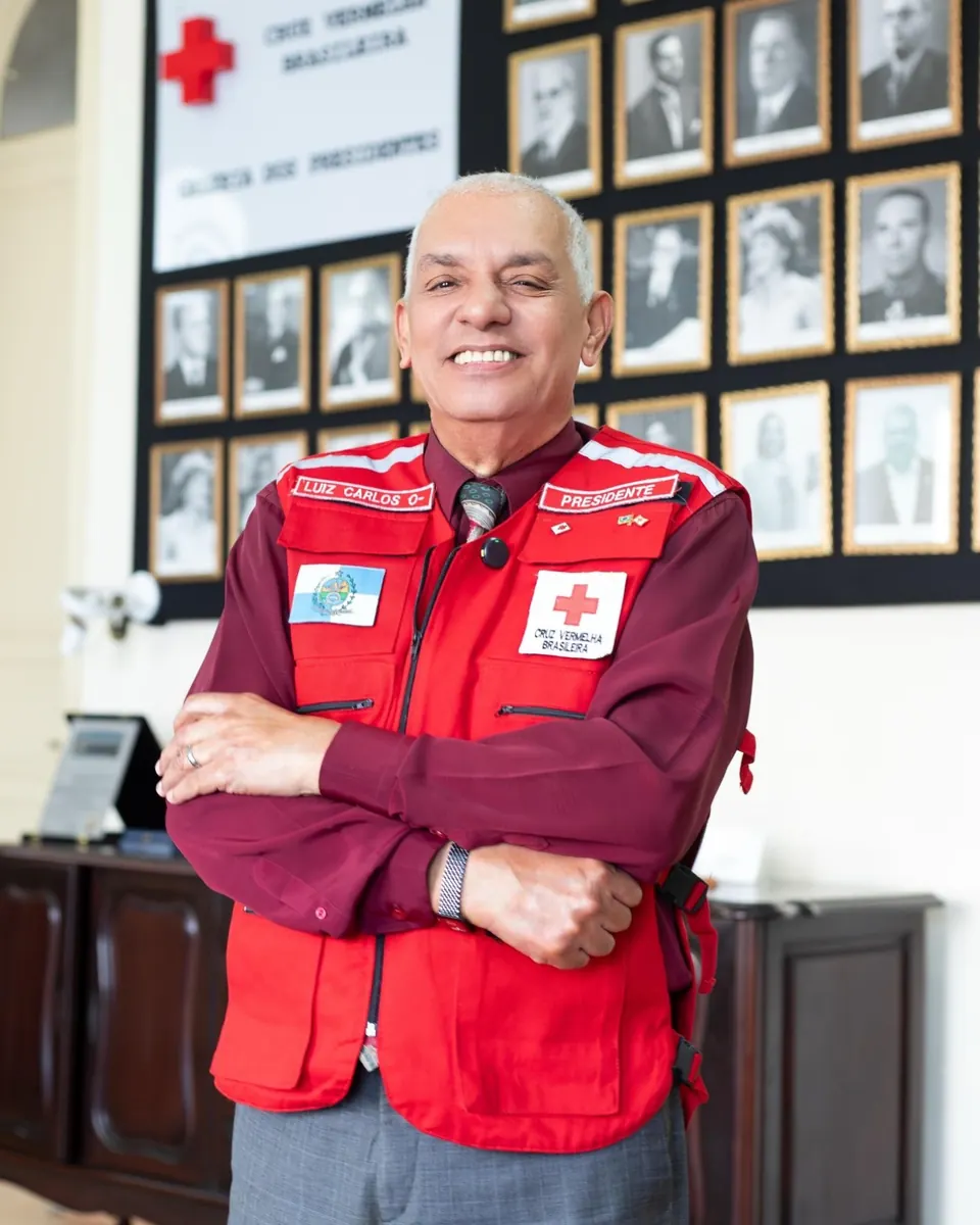 Luiz Carlos dos Santos, presidente da filial, de braços cruzados e sorrindo, com colete vermelho com o emblema e o nome Cruz Vermelha Brasileira, diante dos retratos da Galeria dos Presidentes