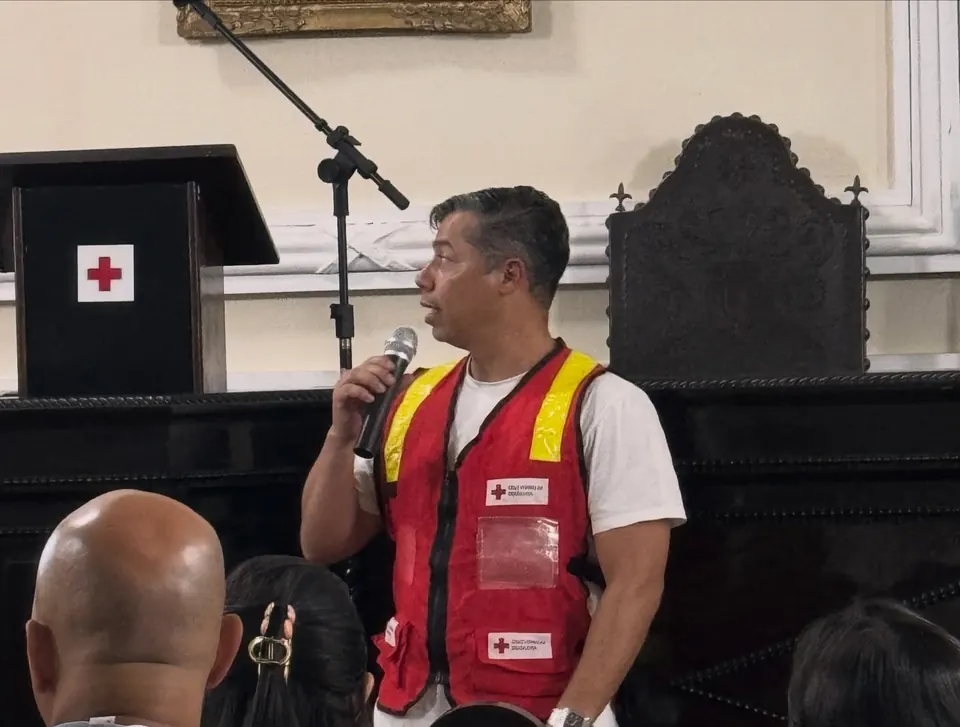 Voluntário da filial, de colete vermelho, fala ao microfone diante da turma, numa capacitação na sede