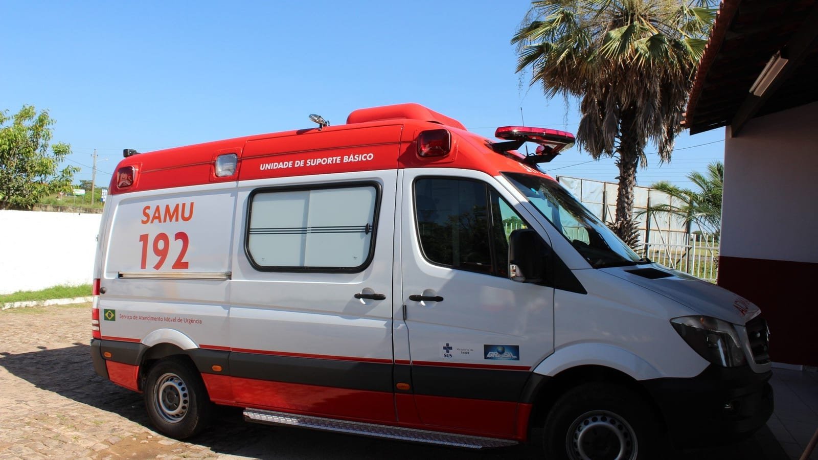 Ambulância branca e vermelha do SAMU 192, unidade de suporte básico, estacionada ao sol ao lado de palmeiras