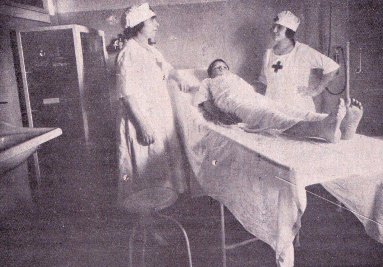 Fotografia antiga em preto e branco de um quarto de hospital, com duas enfermeiras e um paciente no leito