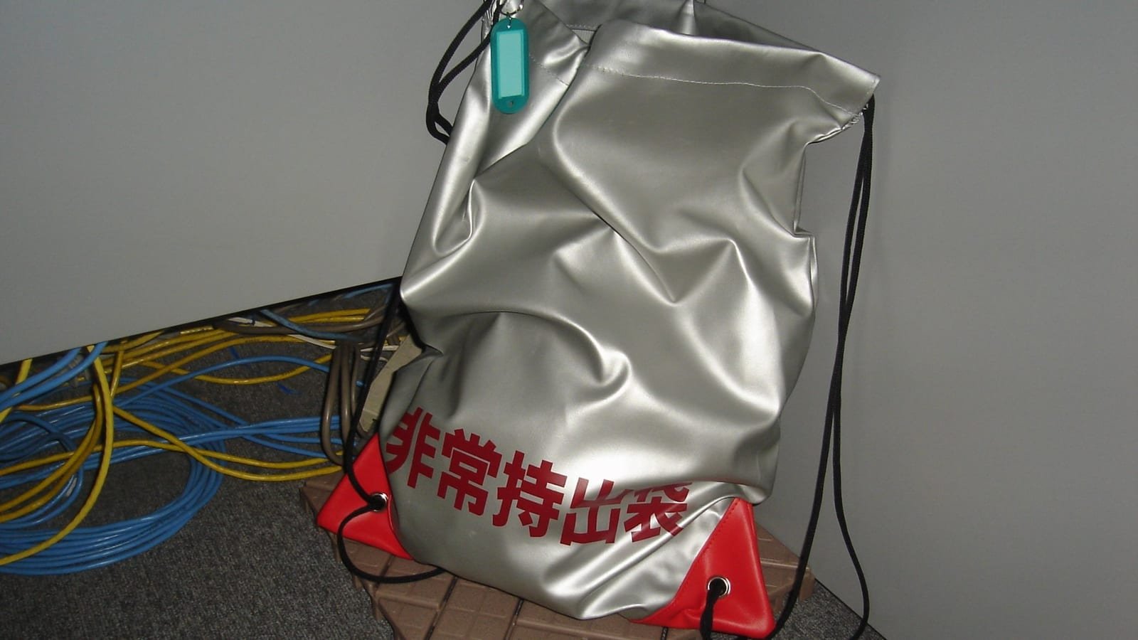 Mochila prateada de emergência, com inscrições em japonês, apoiada no canto de uma sala