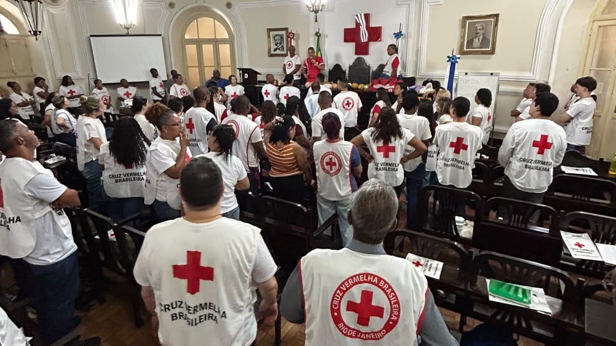 Voluntários de colete e camiseta da Cruz Vermelha, de costas, em um salão de treinamento de primeiros socorros