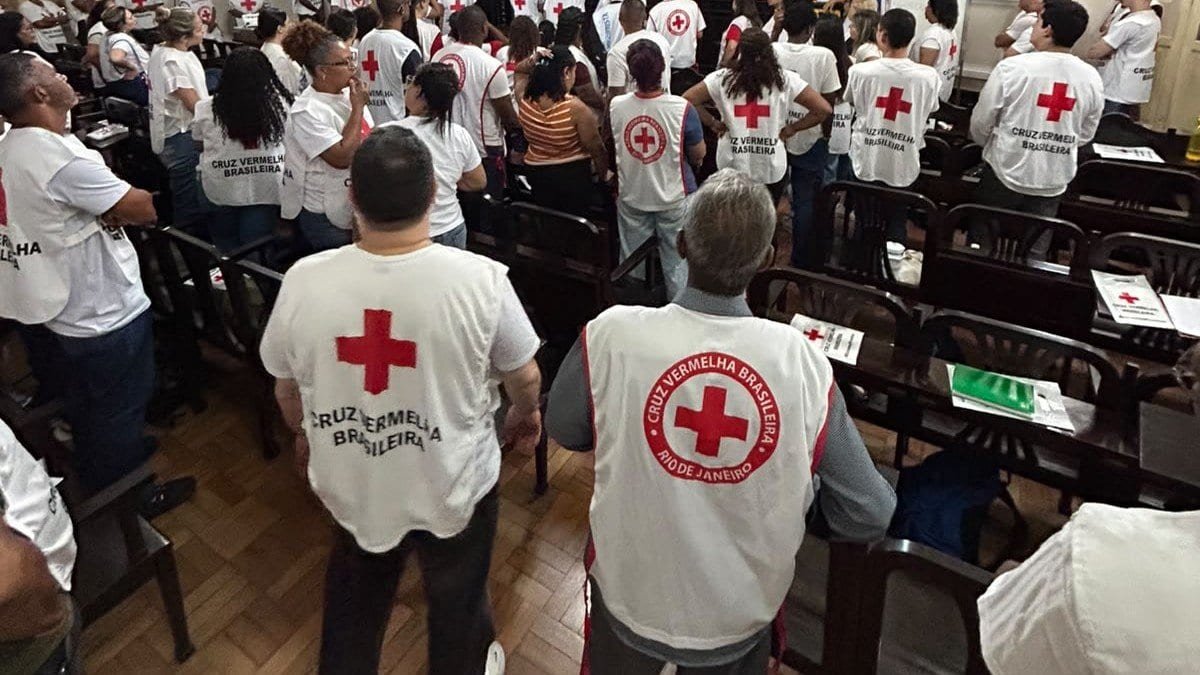 Voluntários de colete branco com a cruz vermelha, vistos de costas, de pé entre fileiras de cadeiras em um salão
